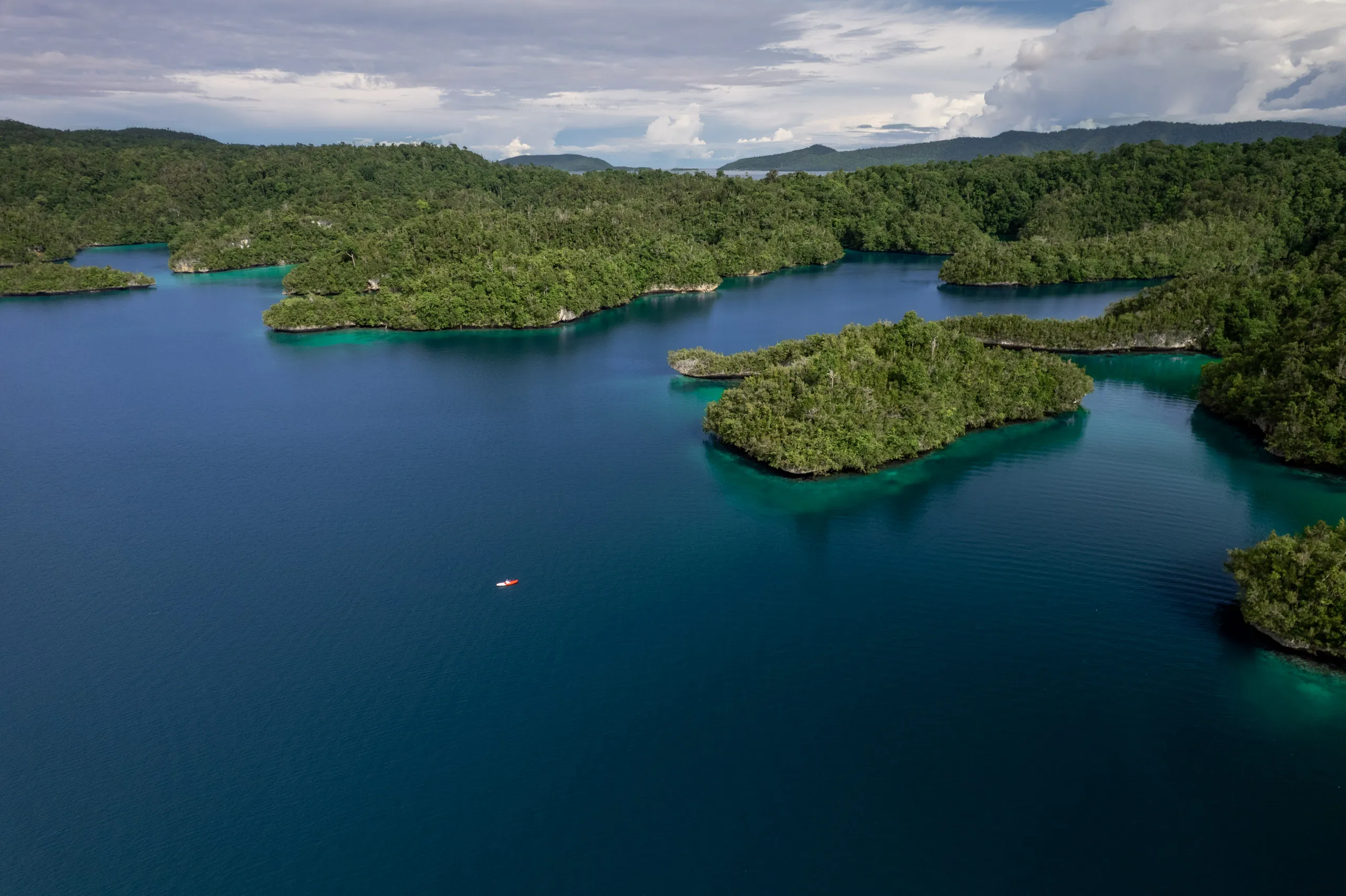 Wide aerial view of a calm bay with a red kayak far out on deep blue water, forested islands along the shore