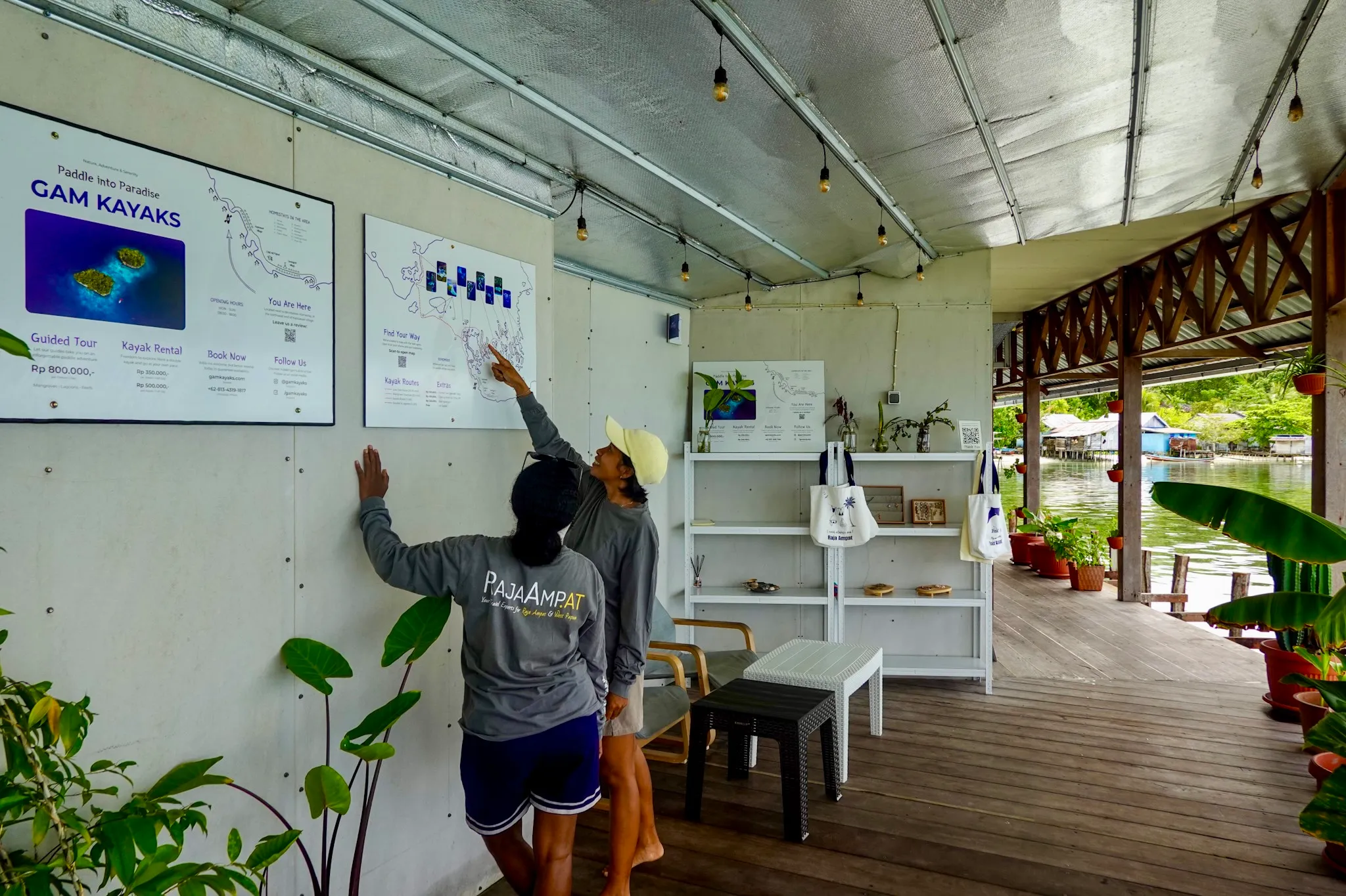 Two people at the Gam Kayaks information wall on the covered base deck, pointing at the route map, the bay behind