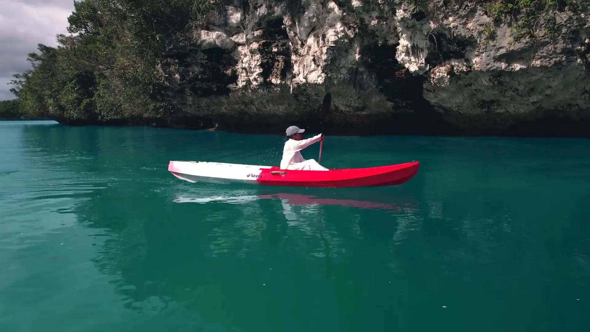 A paddler in a red kayak on emerald water beneath an undercut white limestone cliff