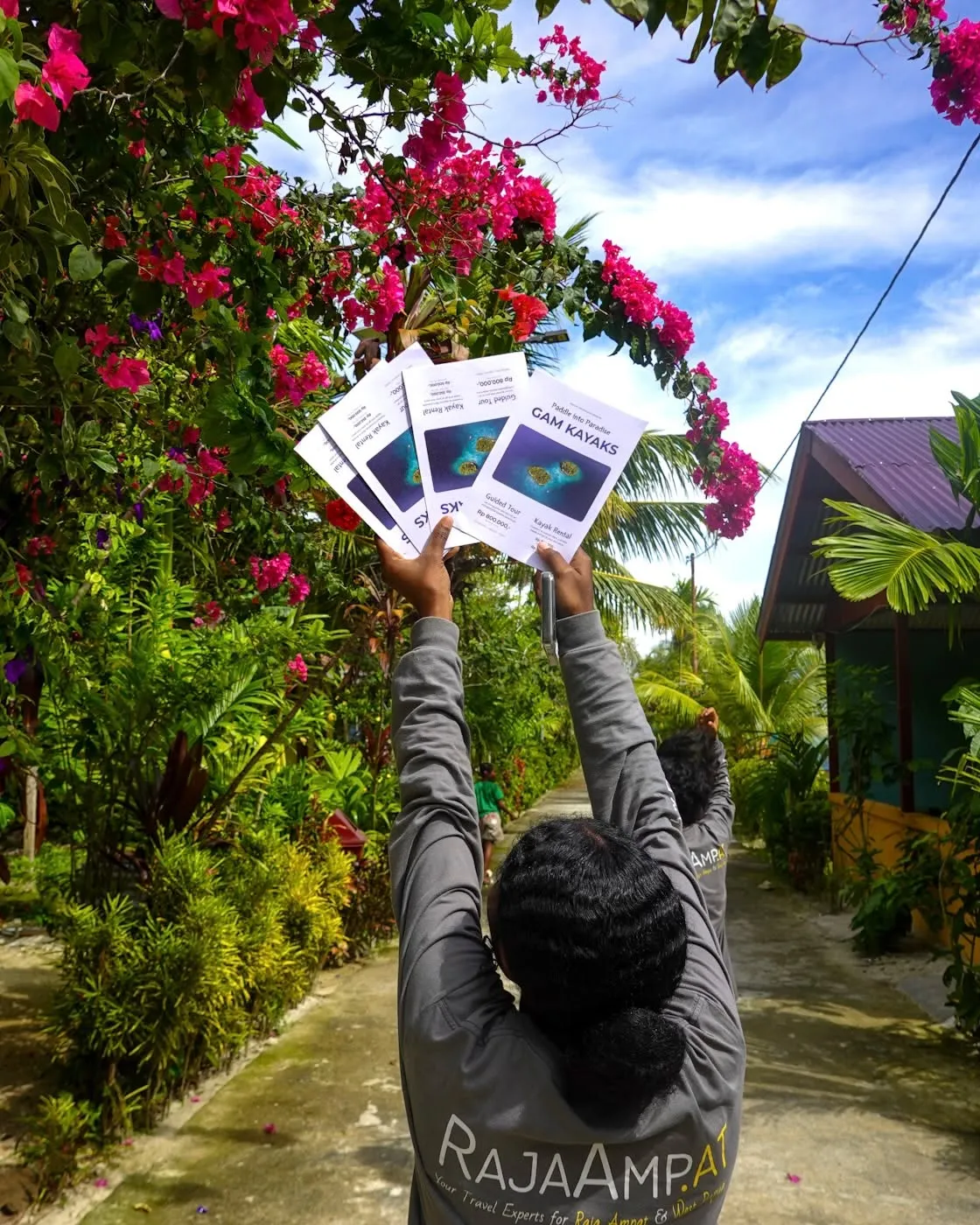 Printed Gam Kayaks price flyers held up against a village path lined with bright pink bougainvillea