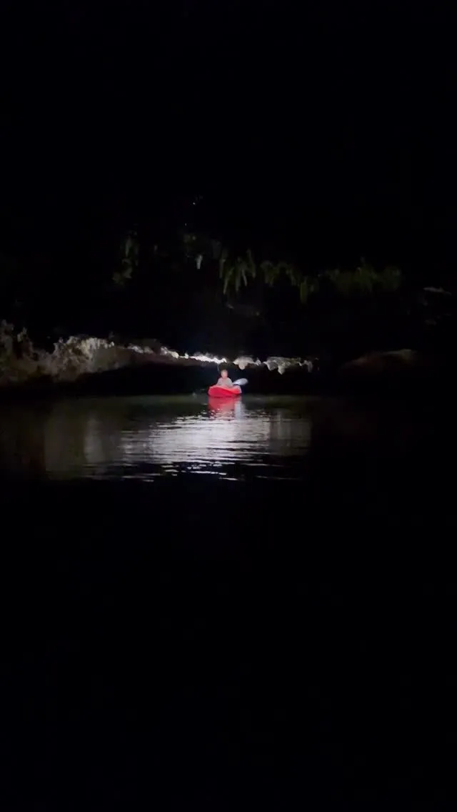 A lit red kayak at night beneath a dark limestone overhang, reflected on still black water
