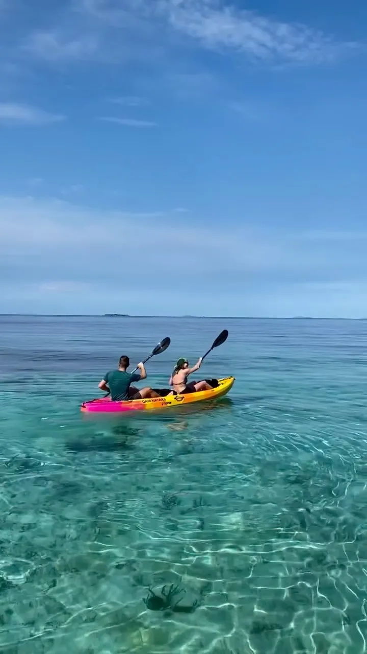 Two paddlers in an orange Gam Kayaks kayak on clear shallow turquoise water, coral visible beneath and a low island on the horizon