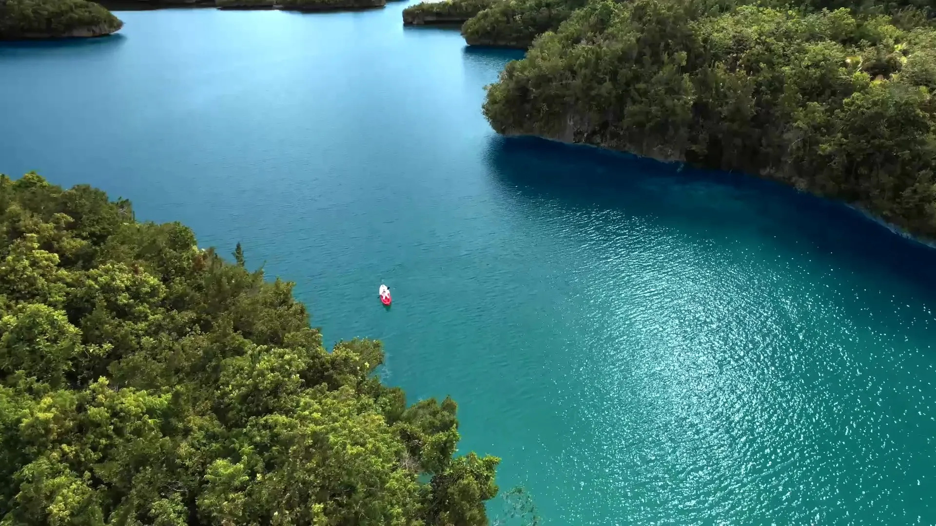 A lone red kayak crossing a deep-blue channel between forested karst islands in Gam Bay