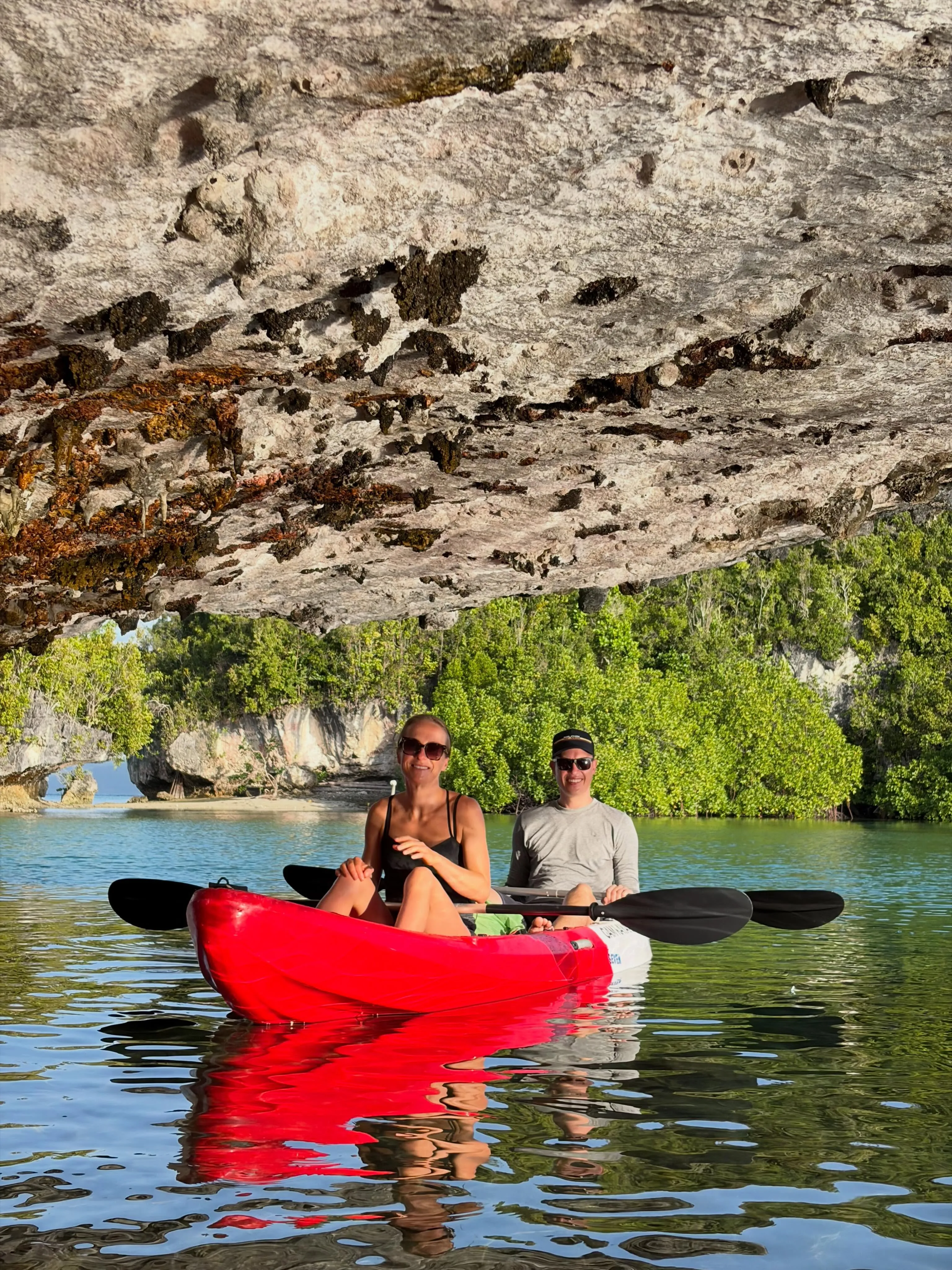 Two paddlers in a red double kayak resting beneath a huge limestone overhang above clear turquoise water