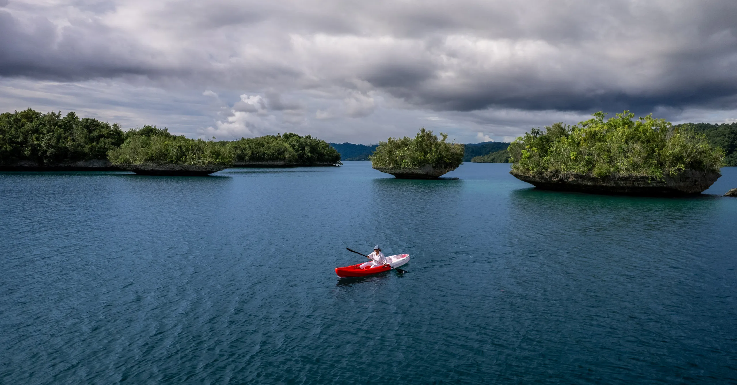 A lone paddler in a red kayak on calm water between small forested karst islets