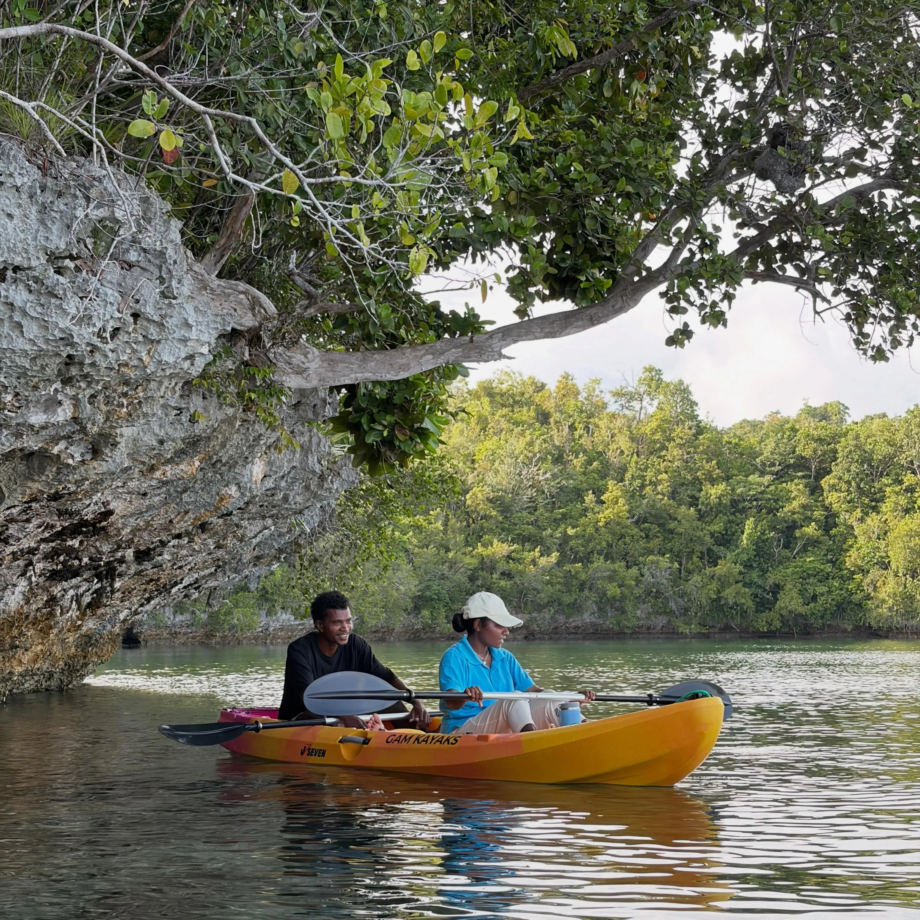 Two paddlers in an orange Gam Kayaks sit-on-top kayak gliding under an overhanging tree beside a limestone cliff on calm green water